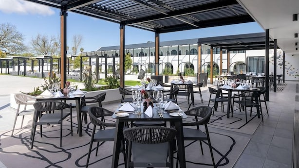 Ein Essbereich im Freien mit Tischen und Stühlen, einer Pergola und Blick auf ein Gebäude mit Bögen im Hintergrund.