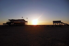Un edificio frente a la playa con una bandera, un sol al fondo y cielo despejado.