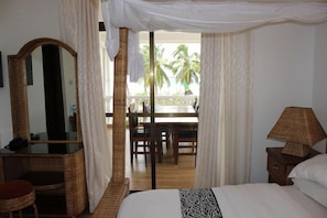 Quarto com cama de dossel, escrivaninha com espelho, luminária e vista para uma varanda com palmeiras.
