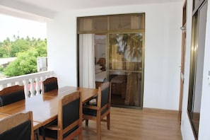 Quarto com mesa e cadeiras de madeira, porta de vidro deslizante para varanda com vista para vegetação.