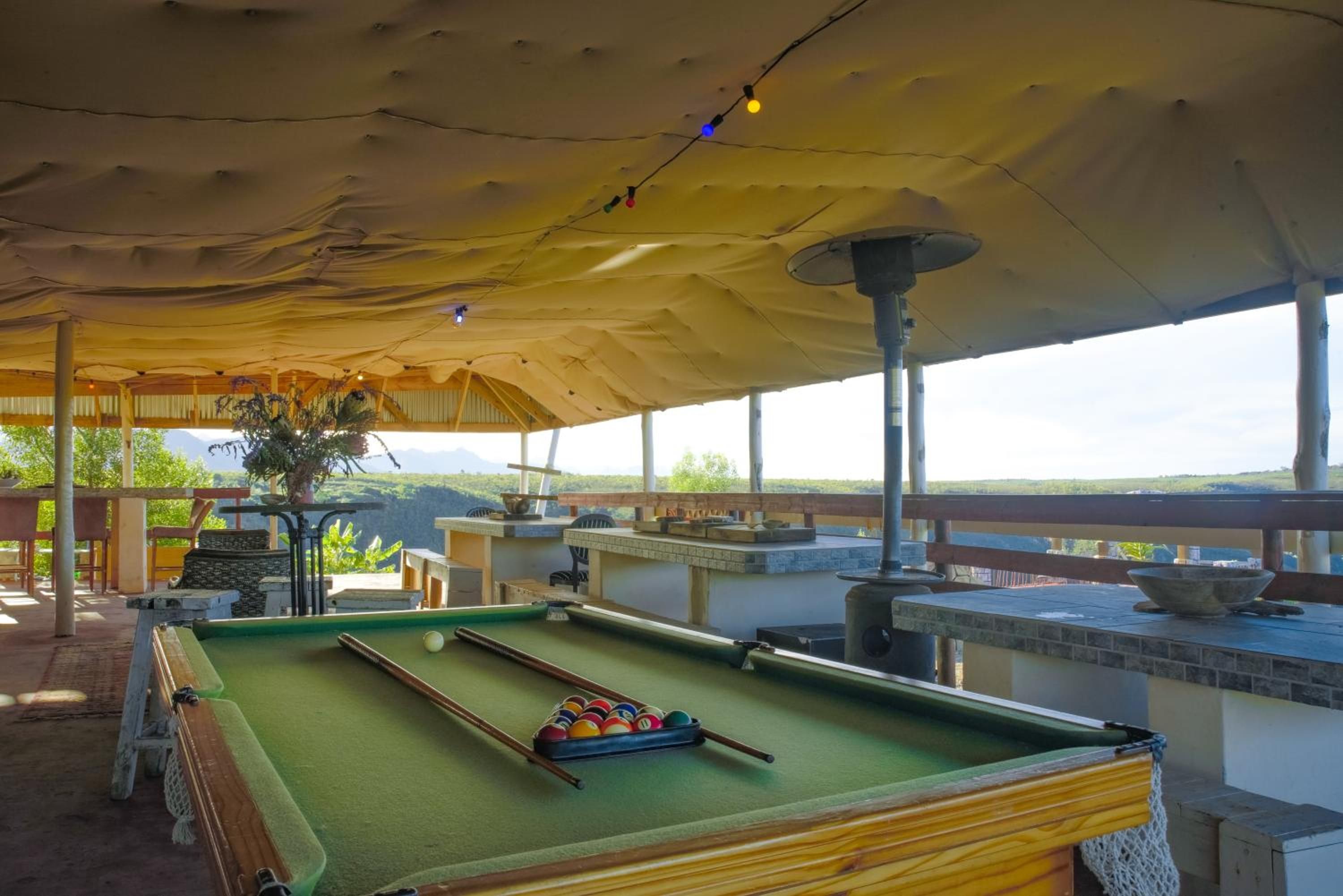 An outdoor pool table with a green felt surface, surrounded by a covered area with a yellow canopy, and a view of the landscape.