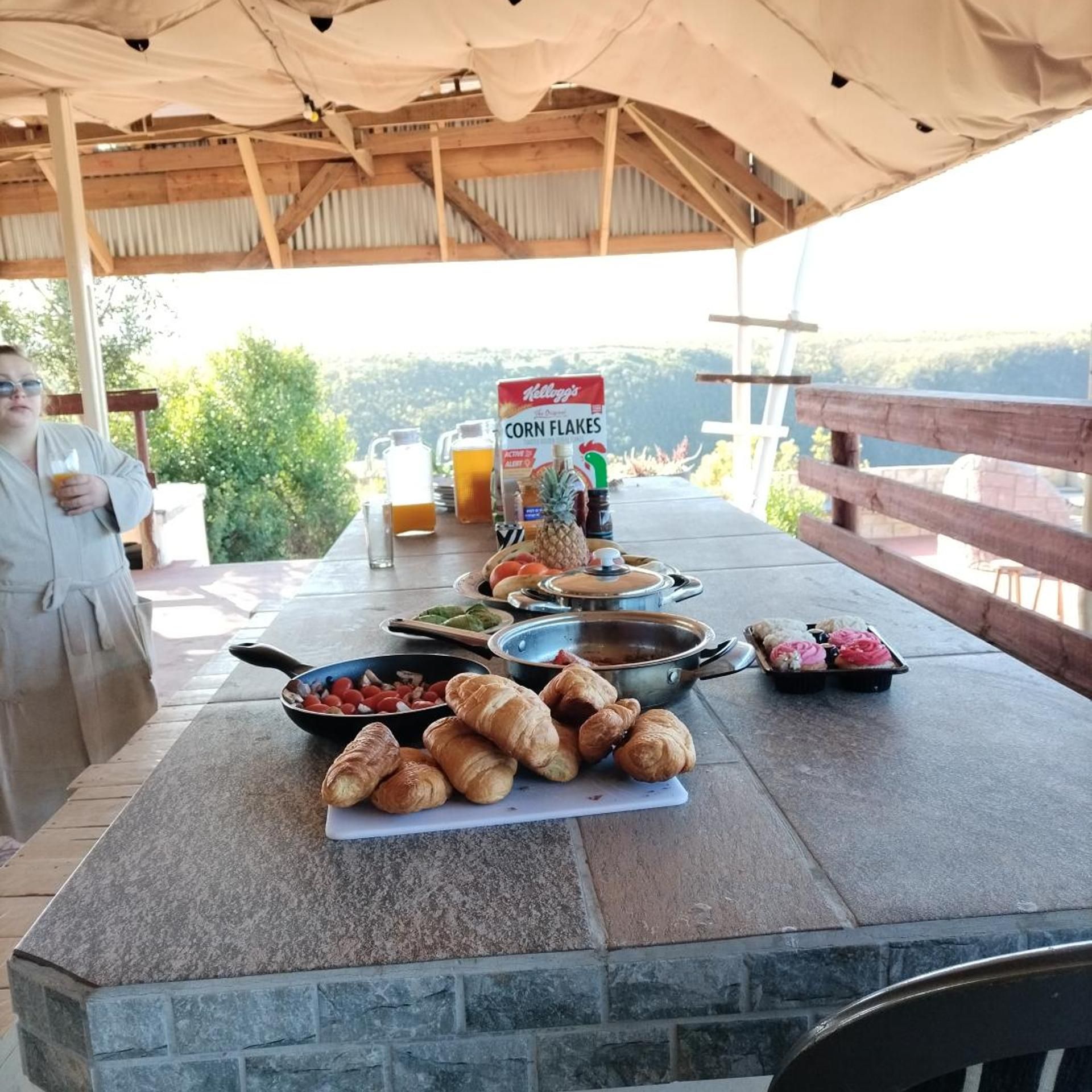 An outdoor breakfast setup with a variety of food items and beverages, including corn flakes, croissants, and a beverage pitcher.