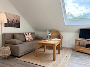 Ein gemütliches Wohnzimmer mit einer Couch, einem Holztisch, einem Korbstuhl, einer TV-Konsole und einem großen Fenster.