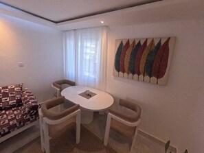 Een knusse, moderne woonkamer met een ronde tafel en twee stoelen, een bank met opvallende kussens en een kleurrijk abstract schilderij aan de muur.