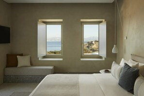 A bedroom with a bed, a bedside table, a lamp, and a view of the sea through two windows. - Parocks Luxury Hotel and Spa (Paros)