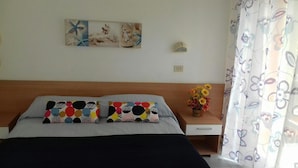 Un dormitorio con una cama, dos almohadas, una mesita de noche con un florero y una lámpara de pared, además de un cuadro.