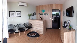 A hotel reception area with a wooden reception desk, three black chairs, a small table with magazines, and a TV mounted on the wall. - Pureza Hotel (Timon)