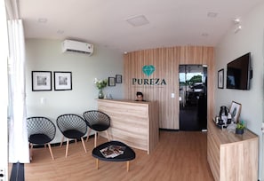 A hotel reception area with a wooden reception desk, three black chairs, a small table with magazines, and a TV mounted on the wall. - Pureza Hotel (Timon)