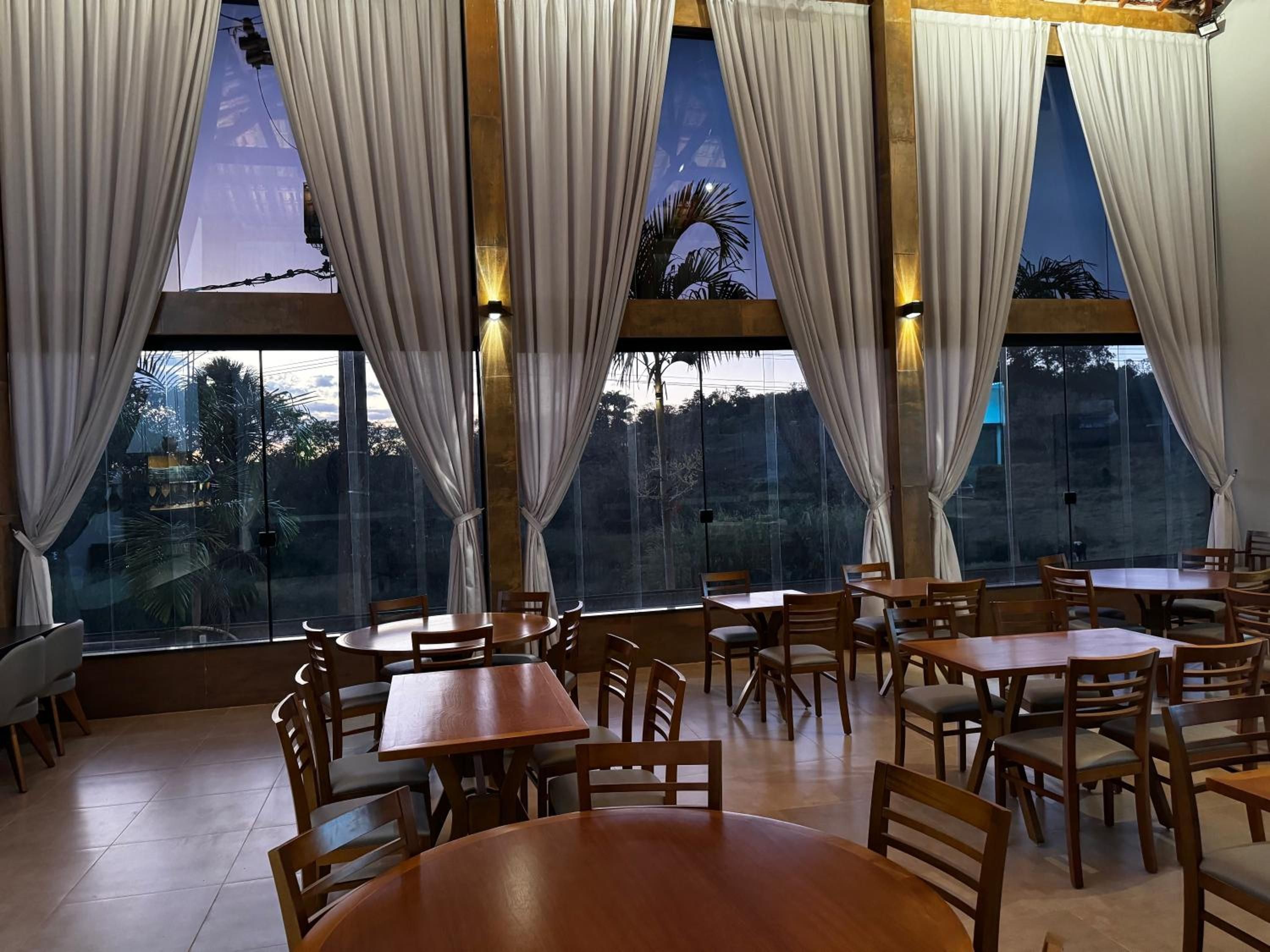 Restaurante com várias mesas e cadeiras, amplas janelas com cortinas e vista para o exterior.