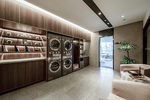 A modern laundry room with built-in washers and dryers, a seating area with a sofa, and a view of the cityscape through a glass door. - Homeinn Plus (Changzhou)
