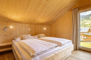 A wooden cabin bedroom with two beds, a window with curtains, and a view of a house and landscape. - Cabin with 1 bedroom (St. Georgen am Kreischberg)