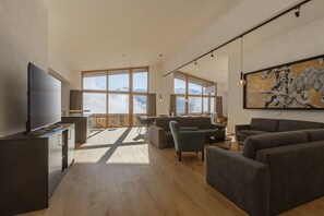 Ein modernes Wohnzimmer mit großem Flachbildfernseher, mehreren Sitzgruppen und großen Fenstern mit Blick auf die Berge.