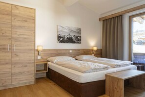 Quarto de hotel com guarda-roupa de madeira, cama com duas almofadas, mesa de cabeceira com abajur e janela com cortinas.
