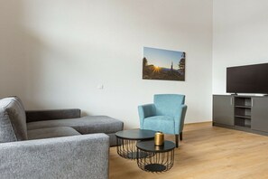 Ein modernes Wohnzimmer mit einem grauen Ecksofa, einem blauen Sessel, einem kleinen runden Couchtisch und einem Flachbildfernseher auf einem Schrank.