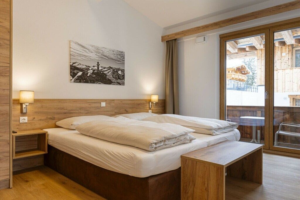 A hotel room with a wooden bed, two bedside lamps, a wooden bench, a large window, and a mountain landscape painting. - Holiday apartment with 3 bedrooms (Mittersill)