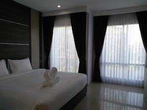 Habitación de hotel con una cama grande, dos ventanas con cortinas traslúcidas y una decoración de toallas con forma de cisne.