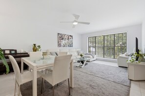 A modern living room with a dining area, a ceiling fan, a large window, and a painting on the wall. - Spacious 3-Bedroom Townhouse Near Terrigal Beach (Terrigal)