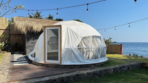 Una tienda en forma de cúpula con puerta de vidrio, rodeada por un muro de piedra, una terraza de madera y una guirnalda de luces.