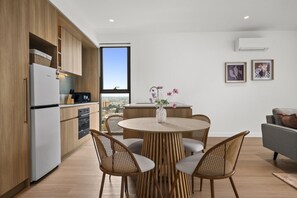 A modern kitchen with a dining area, featuring a round table with chairs, a view through a large window, and a cityscape in the background. - St Kilda Road 2-Bed With Rooftop Access (St Kilda)