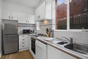 A modern kitchen with white cabinets, stainless steel appliances, and a window overlooking a brick building. - Poolside Art Deco Apartment in St Kilda West (Saint Kilda West)