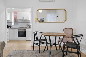A compact kitchen with white cabinets, a stove, and a sink. There is a dining area with a round table and two chairs. A large mirror hangs on the wall. - Poolside Art Deco Apartment in St Kilda West (Saint Kilda West)