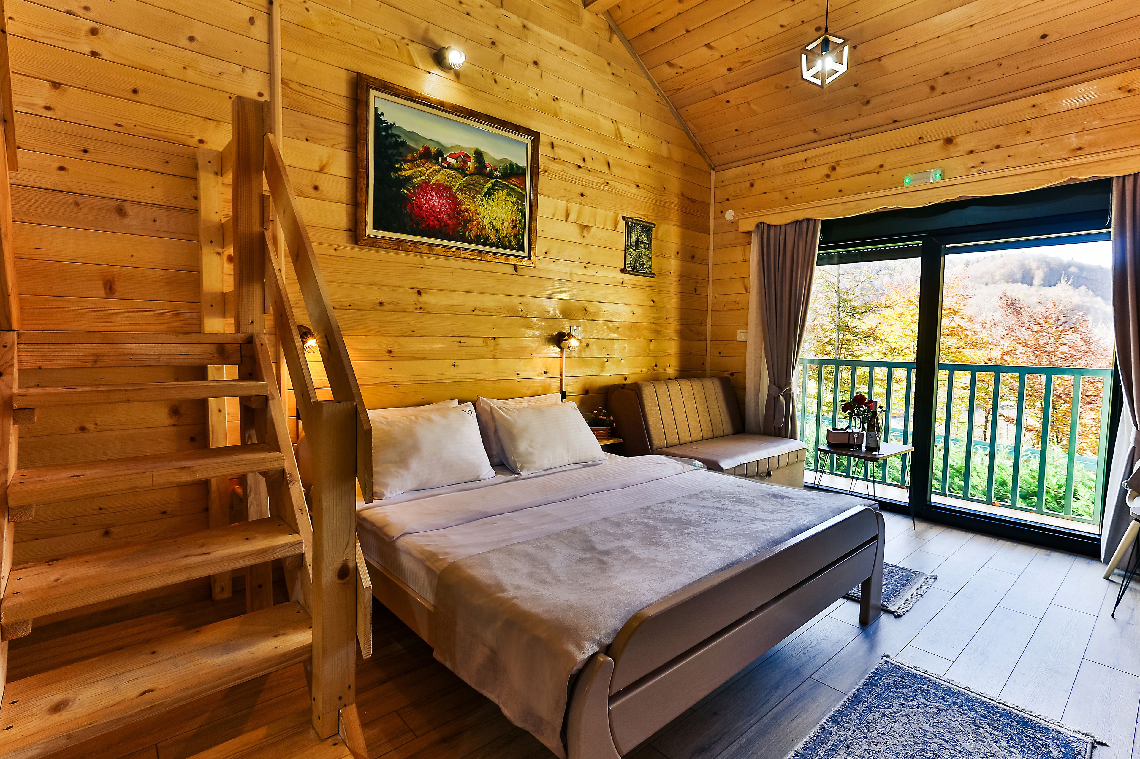A wooden cabin bedroom with a bed, a bench, a ladder, a painting, and a balcony with a view.