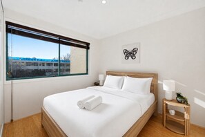 Habitació d'hotel amb un llit gran, una finestra amb persianes, una obra penjada a la paret i una taula de nit amb una làmpada i una planta.