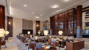 Um saguão de hotel com balcão de recepção, área de estar com mesas e cadeiras, e painéis de madeira decorativos.
