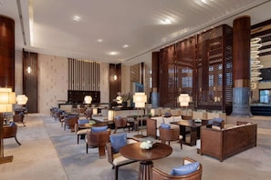 Un vestíbulo de hotel con una recepción, zona de estar con mesas y sillas, y paneles de madera decorativos.