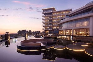 Un hotel moderno con piscina infinita en la azotea, varios balcones e iluminación exterior.