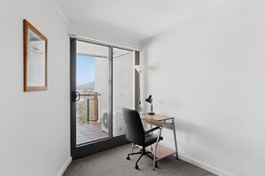 Una habitación pequeña y moderna con puerta de cristal corredera que da a un balcón, un escritorio con una silla y una lámpara de pie.