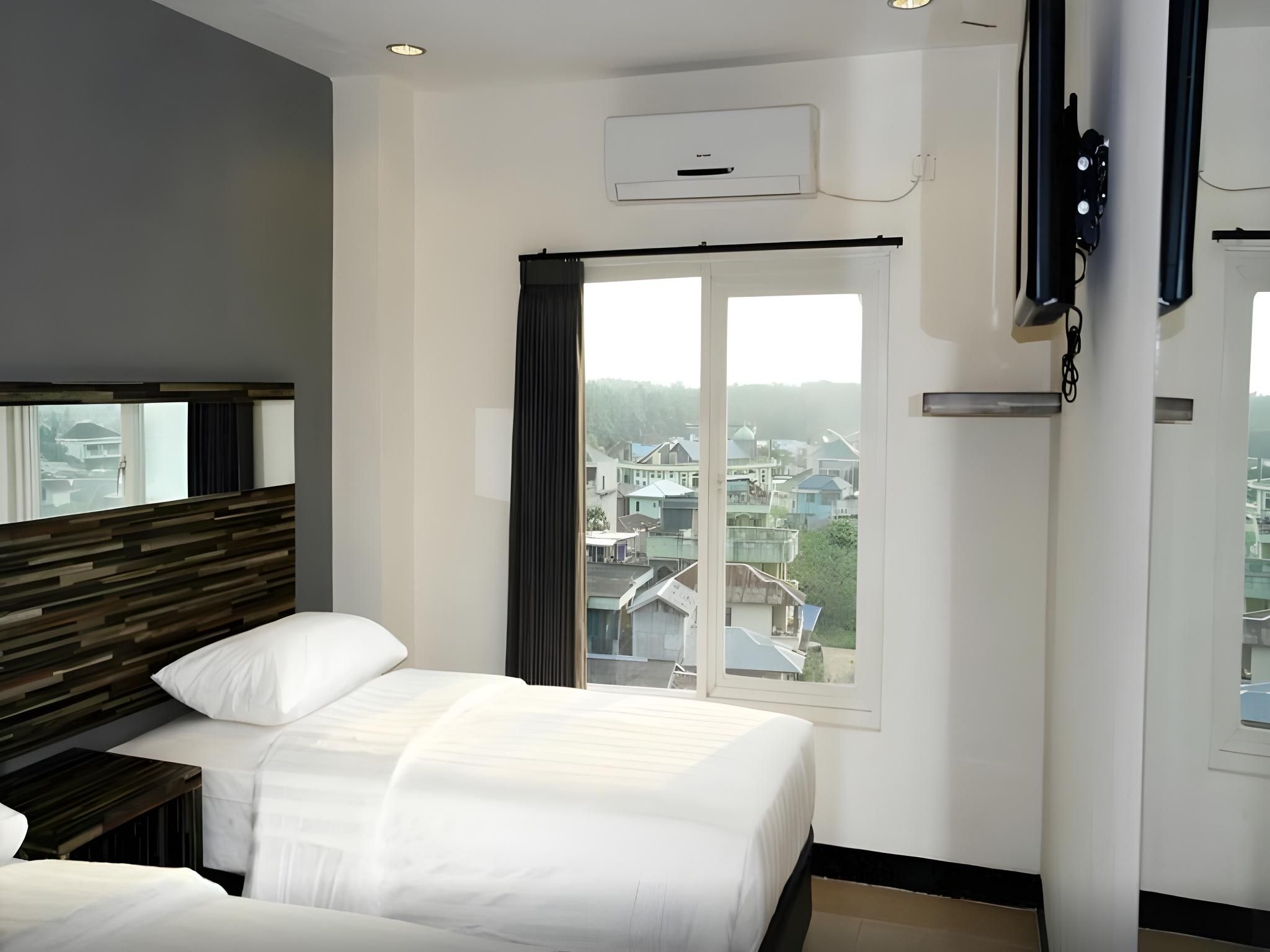 A hotel room with a bed, a nightstand, a window with curtains, an air conditioning unit, and a view of buildings outside.