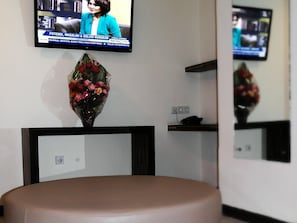 A television mounted on a wall displaying a news program, a decorative vase with flowers, and a brown leather chair. - Galaxy Hotel (Tarakan)