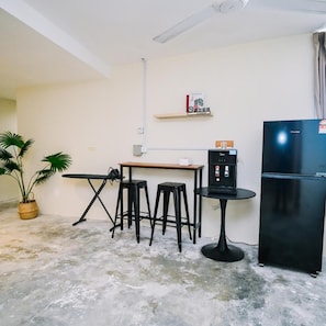 Cozinha compacta com geladeira, cafeteira, mesa, dois banquinhos e um vaso de planta.