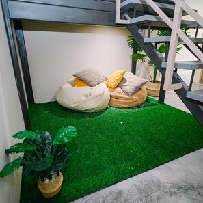 Um espaço de estar pequeno e acolhedor, com chão de grama sintética verde, cercado por uma escada e plantas em vasos.