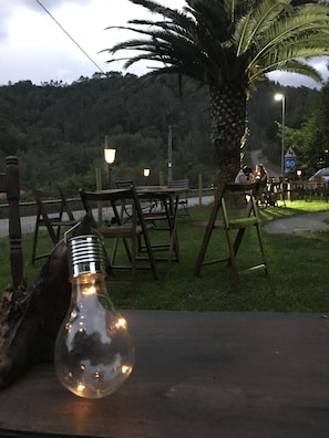 An outdoor seating area with tables and chairs, a light bulb decoration, and a view of a forested hillside. - Hotel Penacabrera 1 (Santa Eulalia de Oscos)