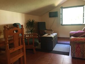 A living room with a wooden chair, a cushioned bench, a potted plant, a television mounted on the wall, and a window providing natural light. - Hotel Penacabrera 1 (Santa Eulalia de Oscos)