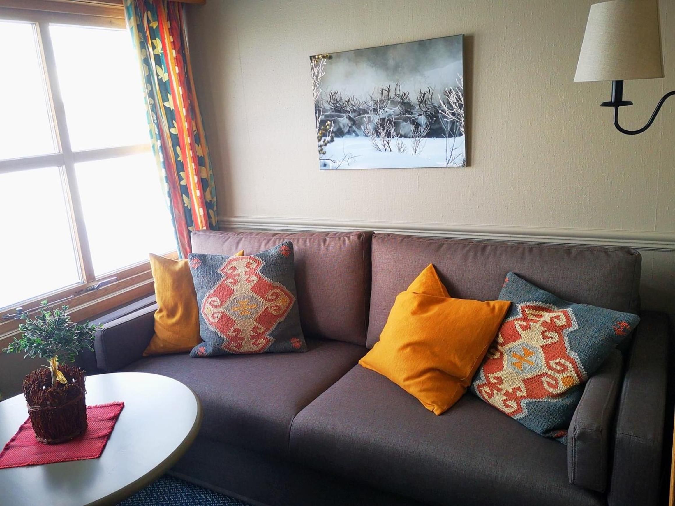 Ein Wohnzimmer mit einer Couch, gemusterten Kissen, einem Gemälde, einer Lampe und einem Fenster mit Vorhängen.