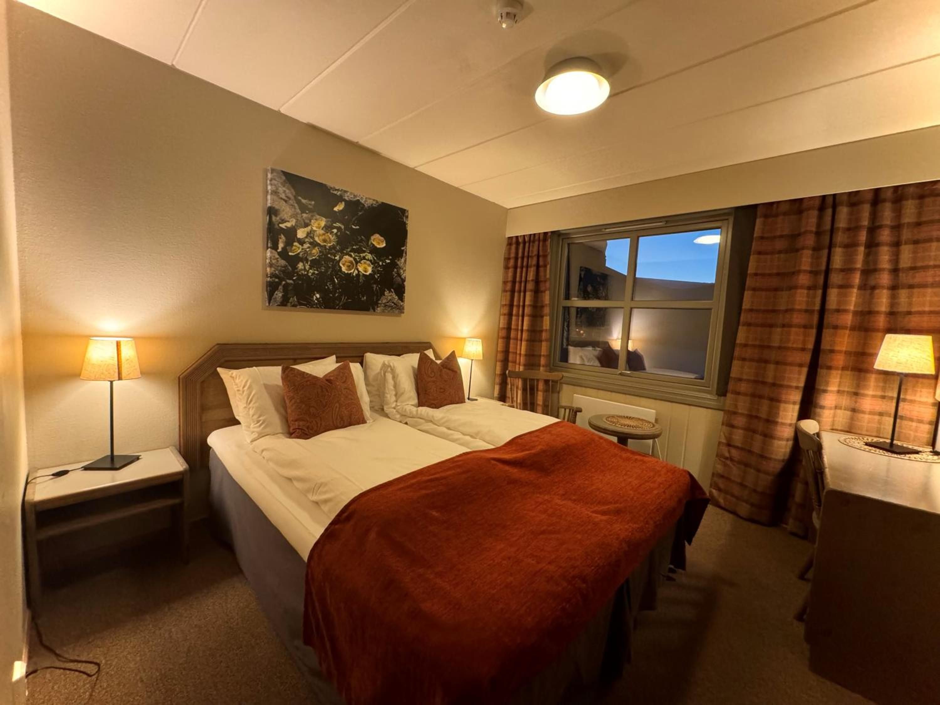 Ein Hotelzimmer mit einem Bett, zwei Nachttischen mit Lampen, einem Fenster mit Vorhängen und einem Gemälde an der Wand.