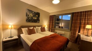 Ein Hotelzimmer mit einem Bett, zwei Nachttischen mit Lampen, einem Fenster mit Vorhängen und einem Gemälde an der Wand.