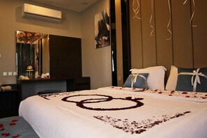 Habitación de hotel con una cama decorada con pétalos de rosa formando un diseño de corazón, un escritorio con lámpara y un aire acondicionado instalado en la pared.