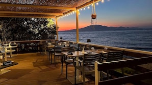An outdoor dining area with tables and chairs, overlooking a body of water and a distant mountain range. - Golden Coast Suites (Zakynthos)