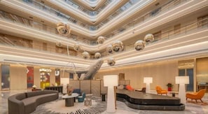 Un hall d’hôtel moderne doté d’un comptoir d’accueil incurvé, d’espaces aménagés avec des sièges et d’un plafond haut éclairé par des luminaires linéaires.