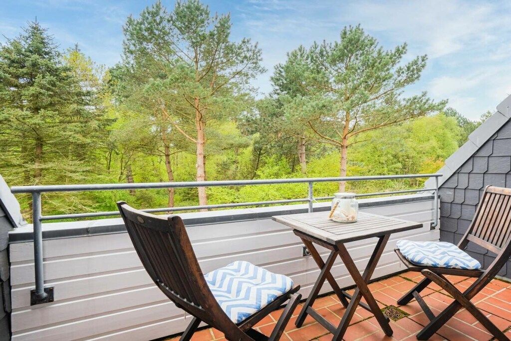 Ein Balkon mit Tisch und zwei Stühlen, umgeben von einem Geländer, mit Blick auf ein bewaldetes Gebiet.
