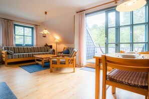 A cozy living room with a sofa, chairs, a coffee table, and a dining table with chairs. There is a large window providing natural light and a view of greenery outside. - OG in the Reimer house (ID 313) (St. Peter-Ording)