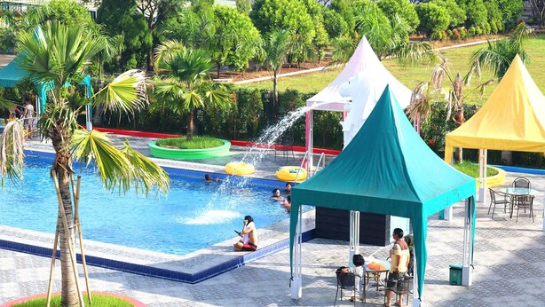 A swimming pool with a water fountain, surrounded by palm trees and covered seating areas. - Singapore City Hotel (Batu Bara)
