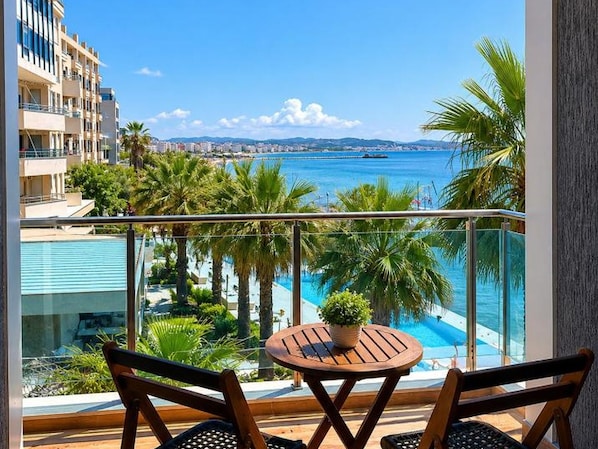 A balcony with a view of a swimming pool, palm trees, and a distant cityscape.