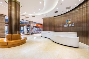 Zona de recepción de hotel con un mostrador curvo, un área de descanso con una columna alta y una pared de madera con señalización.