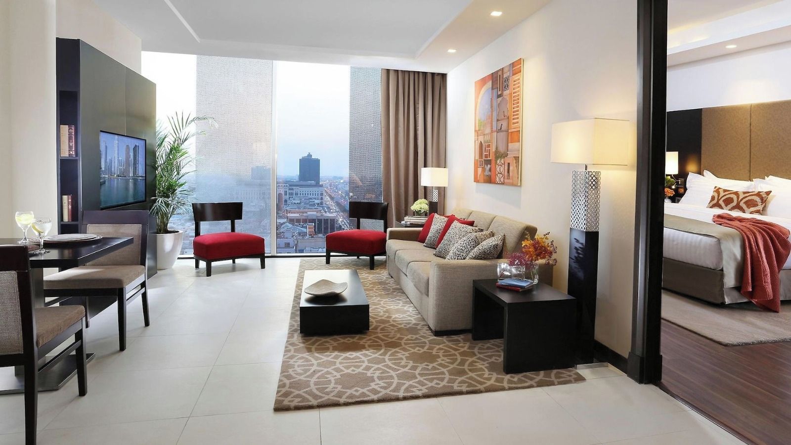 A modern hotel room with a city view, a dining area, and a living space with a sofa and chairs.