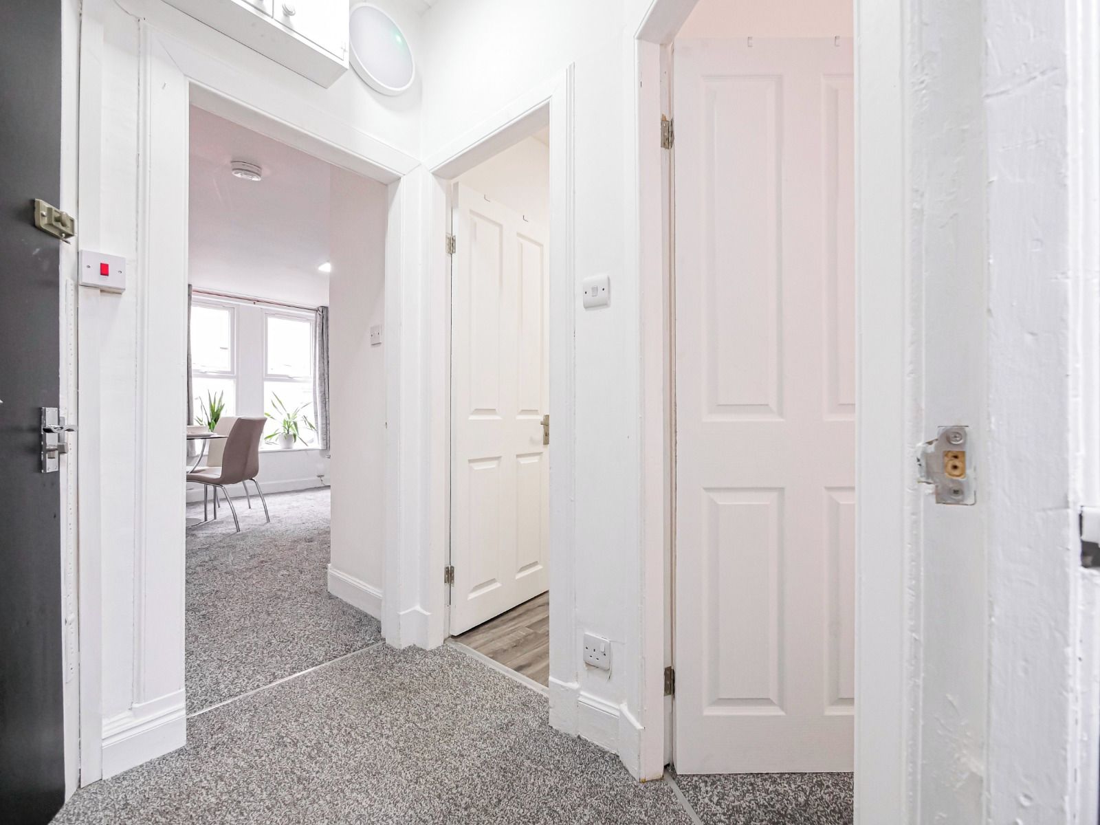 Un pasillo con puertas blancas que dan a una habitación con una mesa de comedor y sillas, piso alfombrado y una ventana con cortinas.